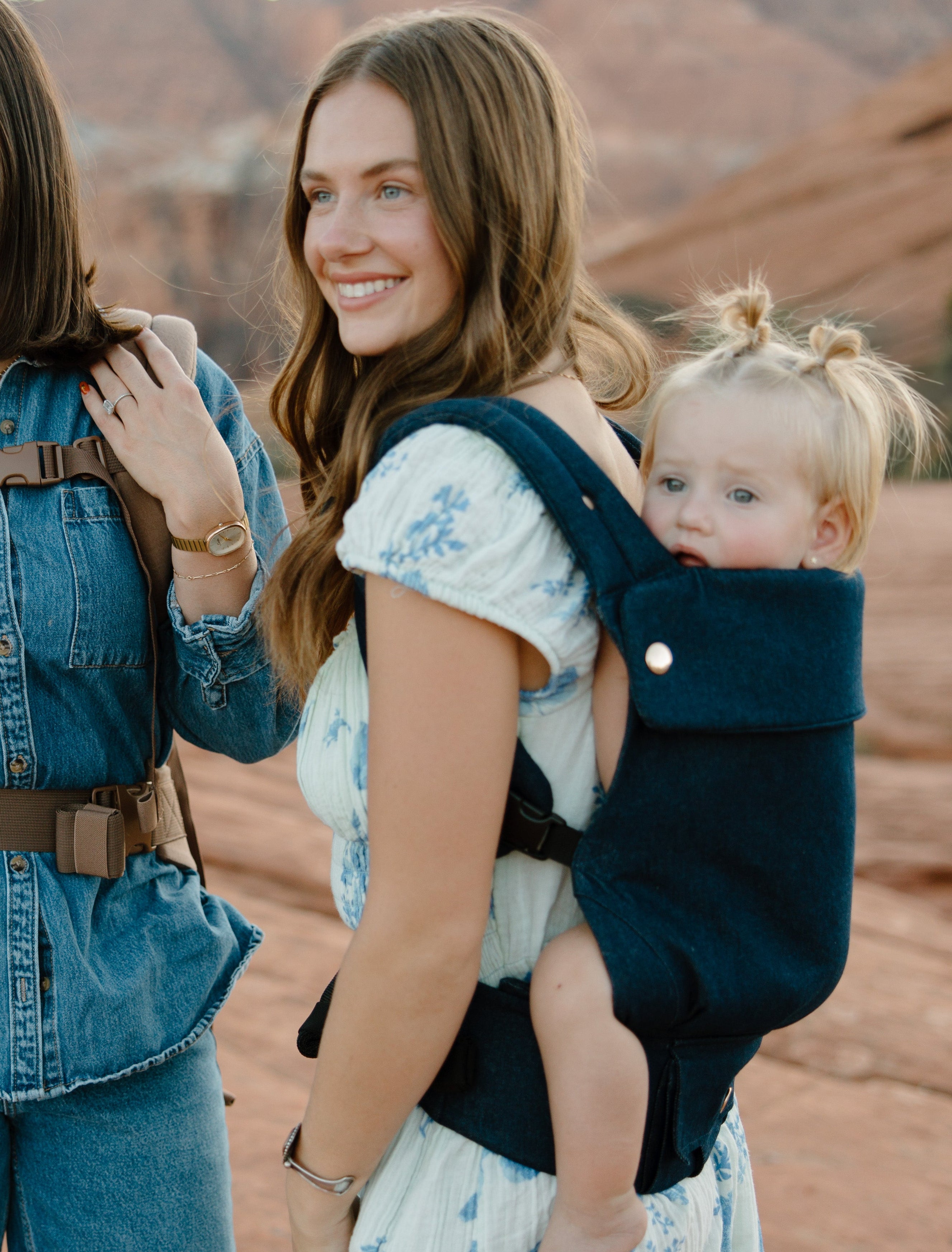 Two women standing in a desert landscape with mountains, one carrying a child in a sling.