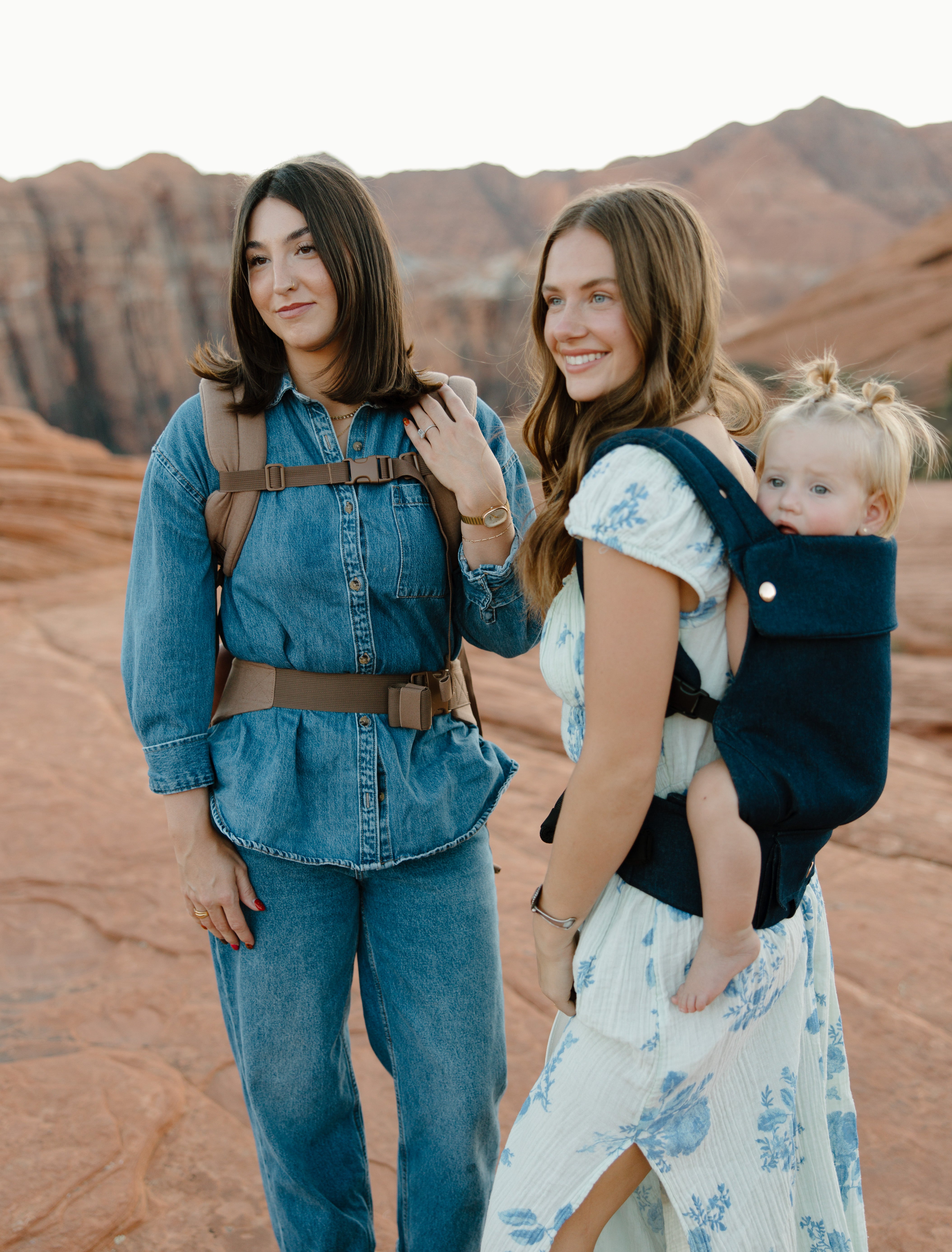 Two women standing in a desert landscape with mountains, one carrying a child in a sling.