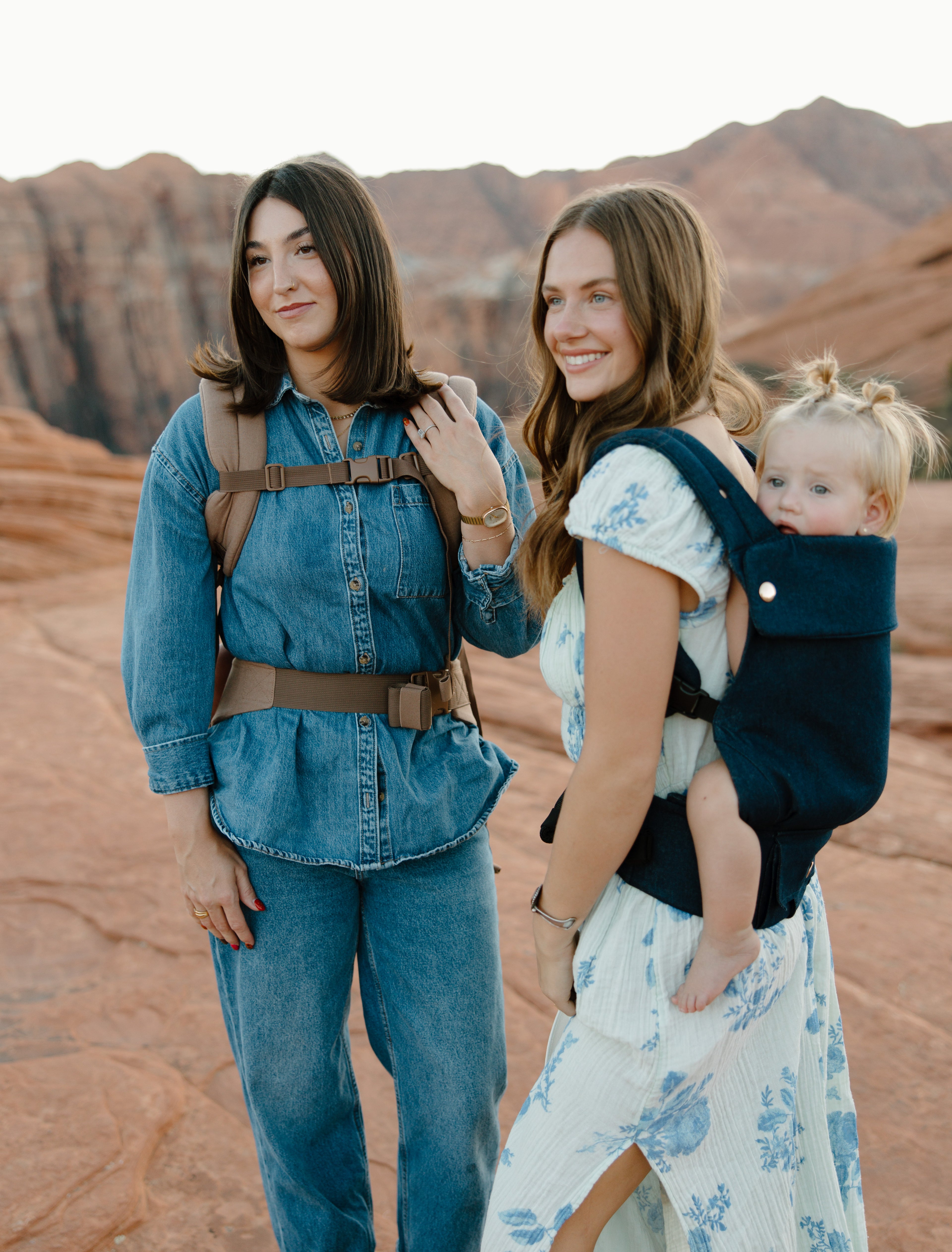 Two women standing in a desert landscape with mountains, one carrying a child in a sling.
